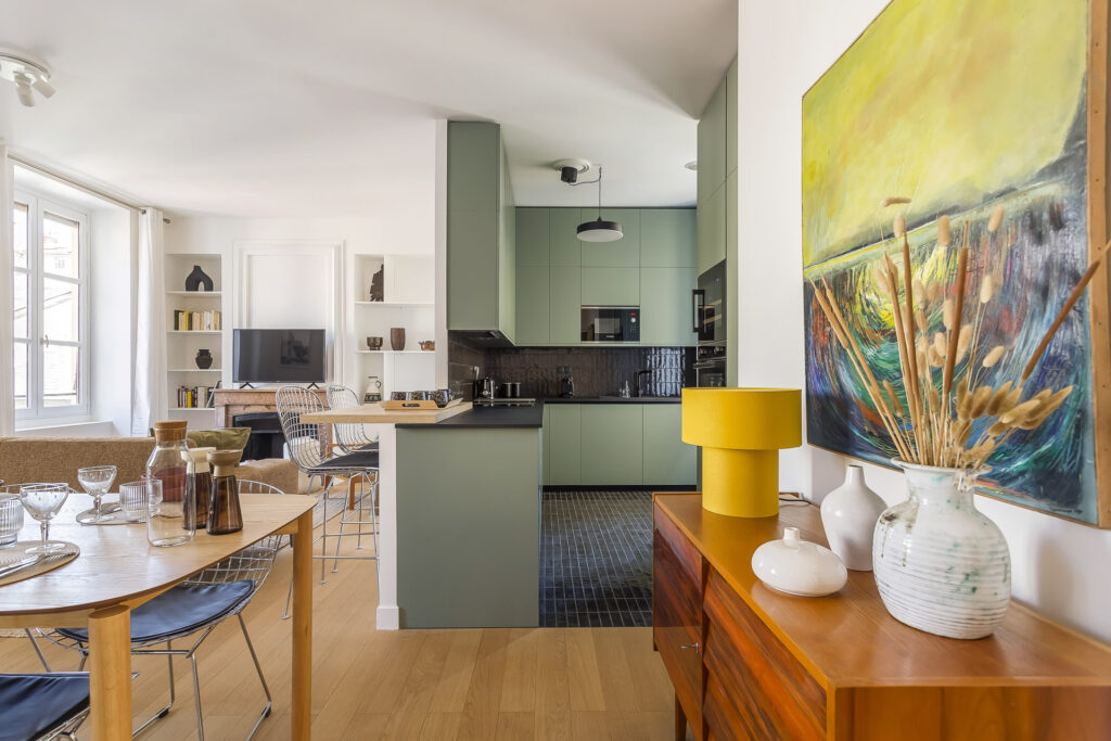 Cuisine et salle à manger modernes et ouvertes avec des meubles vert pastel, un dosseret noir, une table en bois pour les repas - parfait pour les propriétaires exigeants cherchant une gestion locative de courte durée - des œuvres d'art colorées, des vases et de la lumière naturelle dans tout le bâtiment. | 2BeApart Lyon