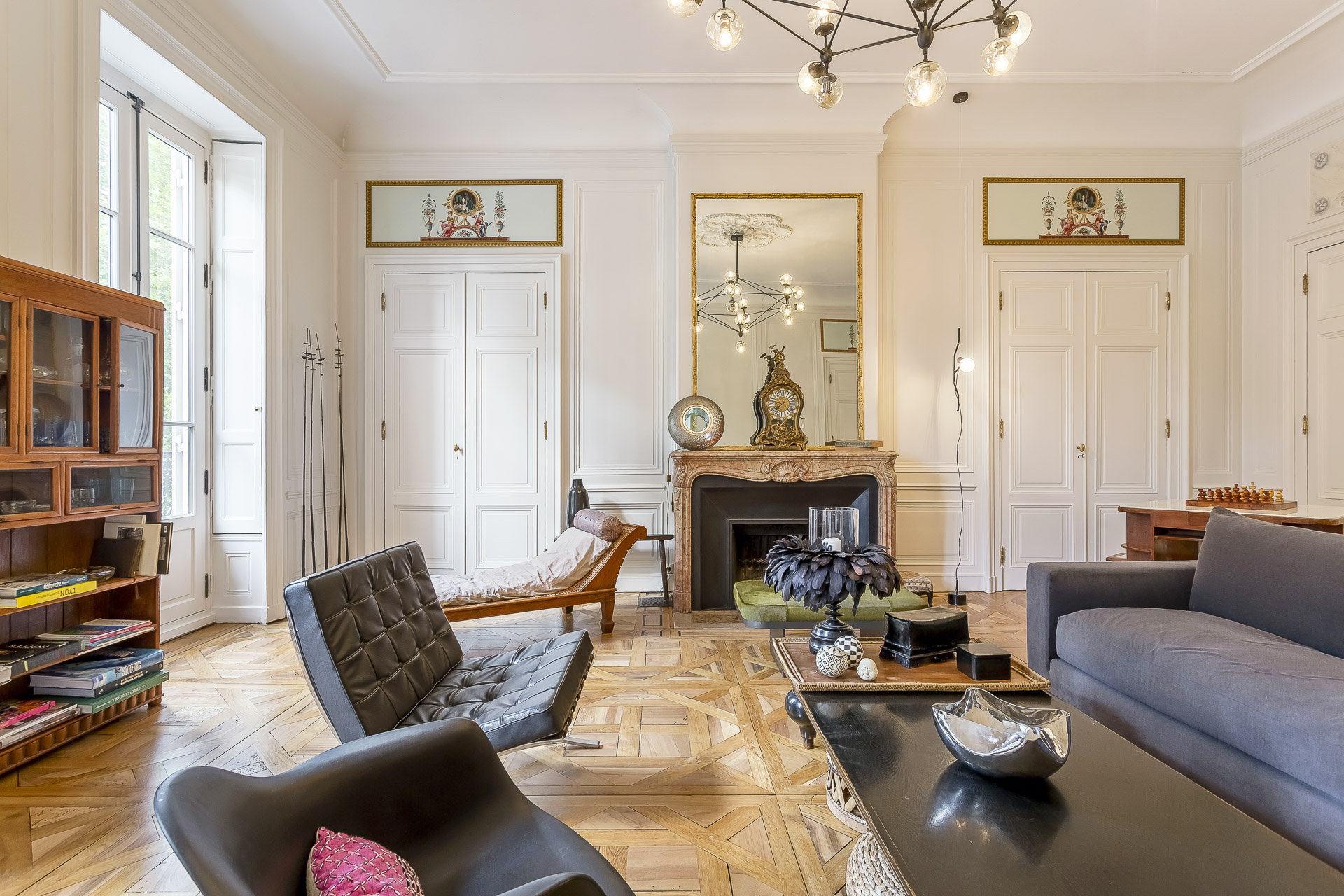 Salon élégant avec parquet, murs blancs ornés, grandes fenêtres, cheminée en marbre surmontée d'un miroir, et mobilier moderne et classique. Parfait pour les propriétaires exigeants à la recherche d'une conciergerie haut de gamme et d'une lumière naturelle dans tout l'espace. | 2BeApart Lyon