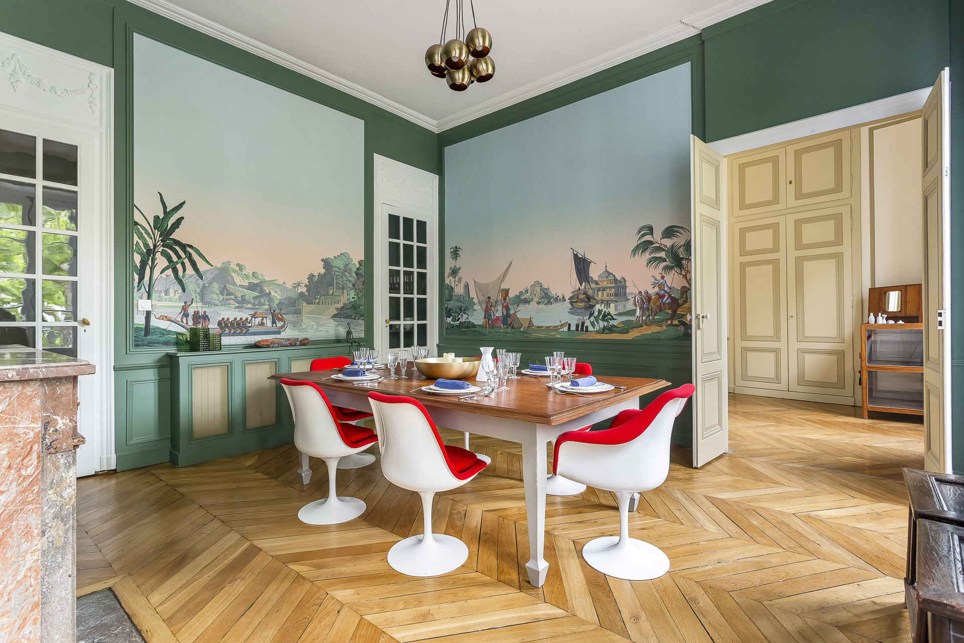 Une salle à manger élégante avec des murs verts, une table en bois pour six personnes, des chaises blanches modernes avec des coussins rouges et de grandes peintures murales représentant des paysages tropicaux et des voiliers - parfait pour une location meublée Lyon ou une conciergerie haut de gamme. | 2BeApart Lyon