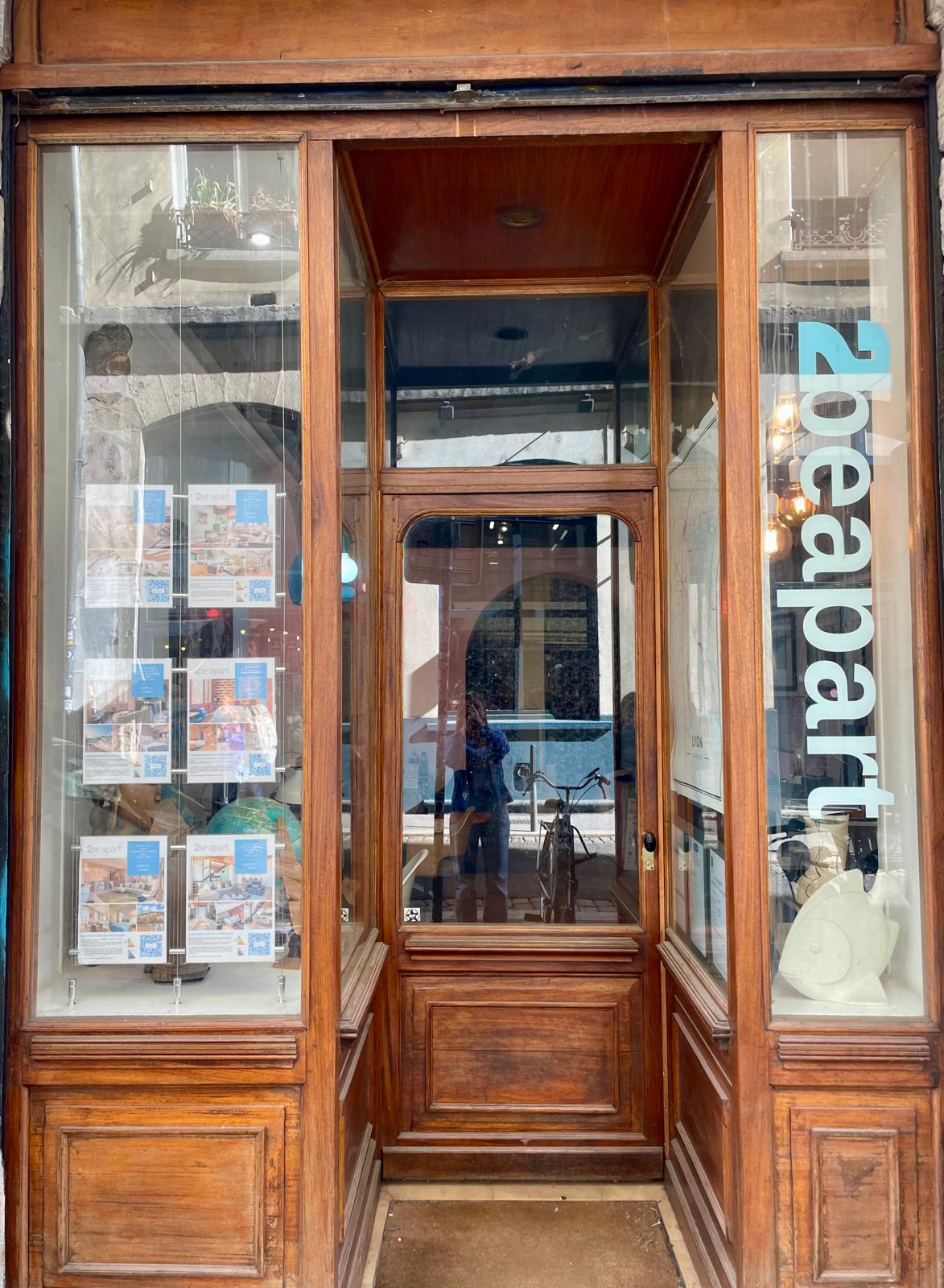Une vitrine en verre à cadre en bois présente des documents imprimés dans la fenêtre, avec "2beapart" écrit verticalement en grandes lettres blanches et bleues. À l'intérieur de l'entrée, un vélo laisse entrevoir un espace conçu pour les propriétaires exigeants ou les services de gestion locative. | 2BeApart Lyon