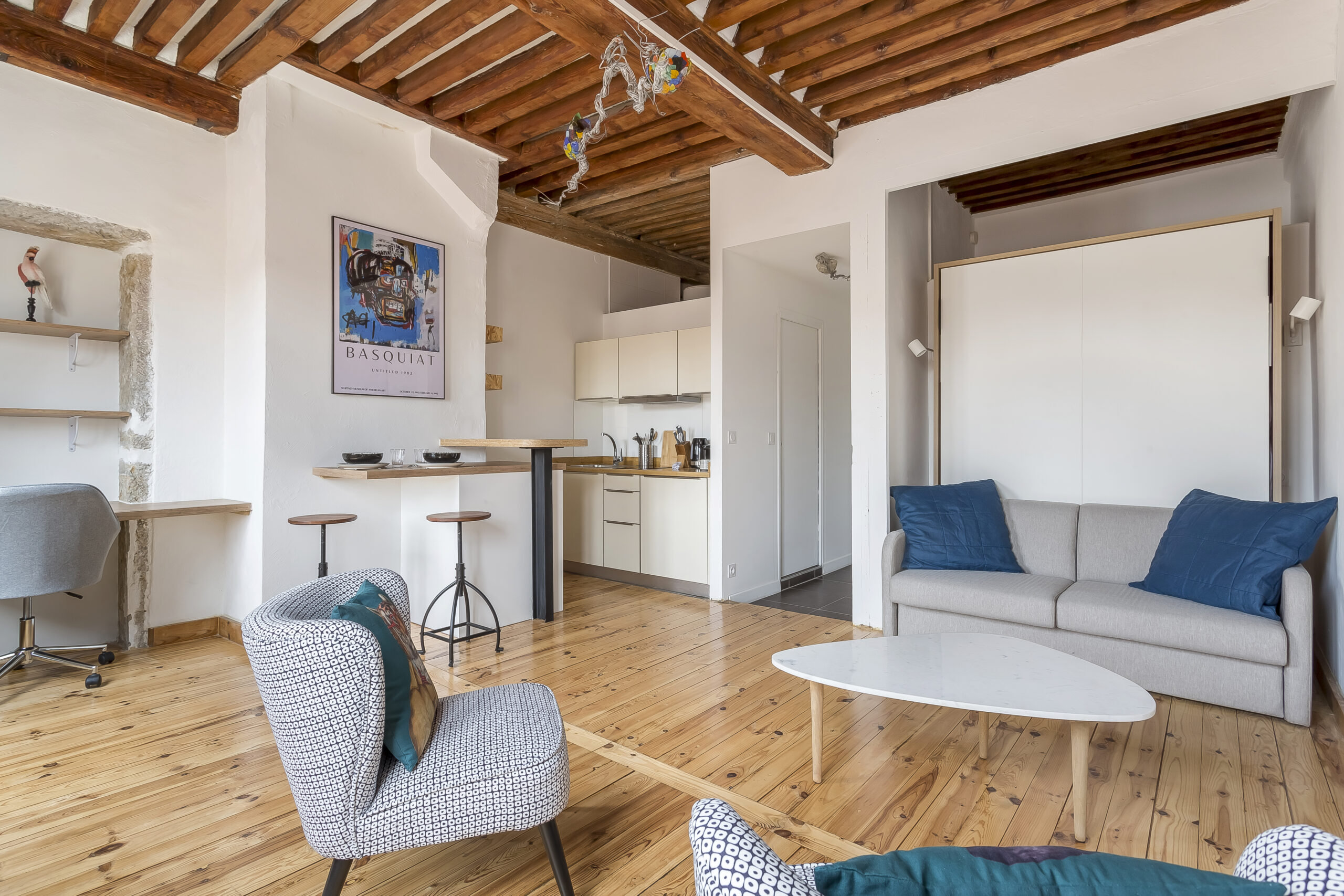 Regulations 7 Un espace de vie moderne et lumineux avec des planchers en bois et des poutres apparentes, comprenant un canapé gris, un fauteuil à motifs, une table basse blanche, un petit coin cuisine et un bar à petit-déjeuner avec deux tabourets. | 2BeApart Lyon