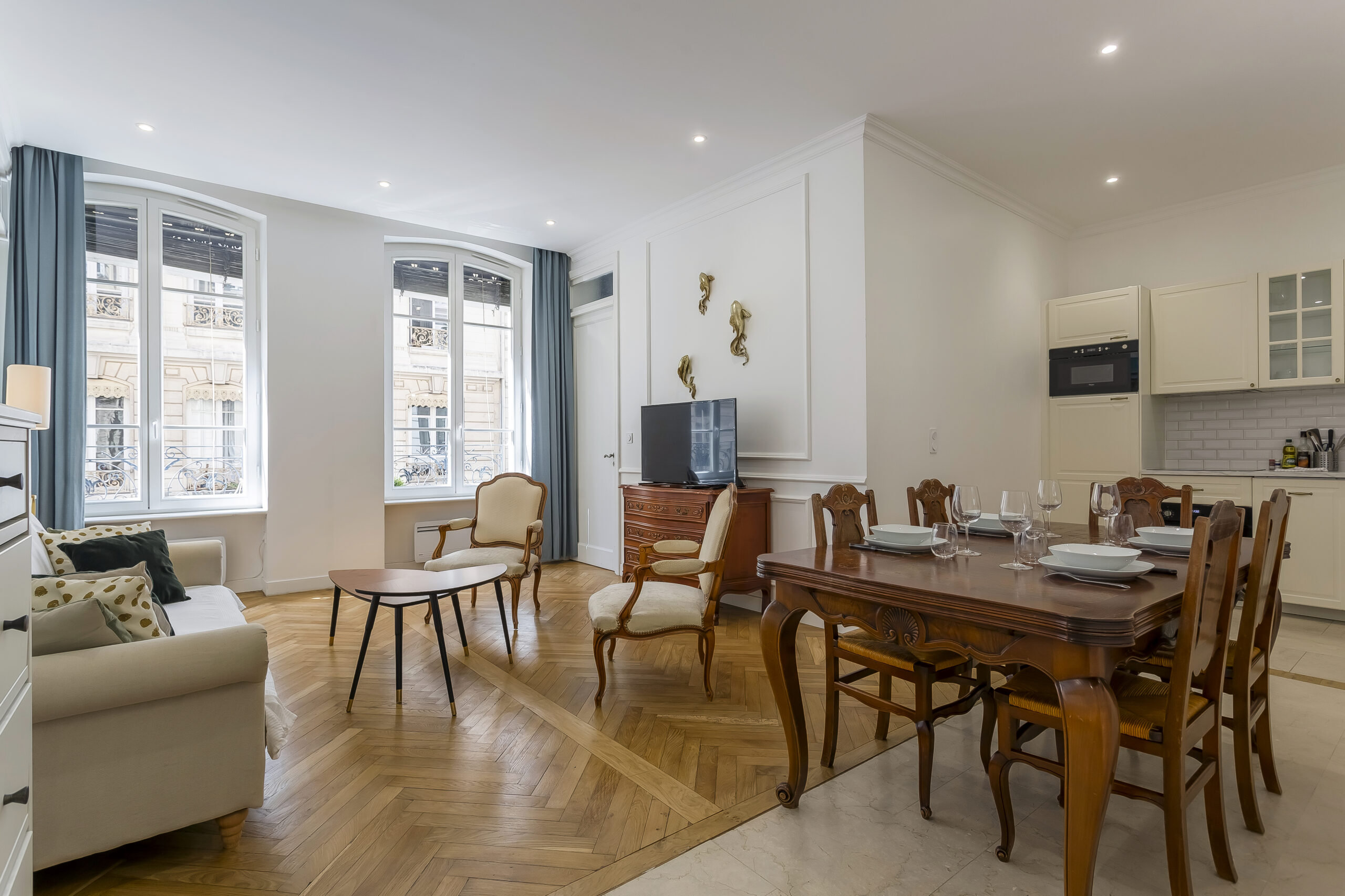 Salon et salle à manger clairs et élégants avec sols en bois, canapé, tables basses, table à manger pour quatre personnes, télévision et cuisine ouverte. De grandes fenêtres avec des rideaux bleus laissent entrer la lumière naturelle. | 2BeApart Lyon