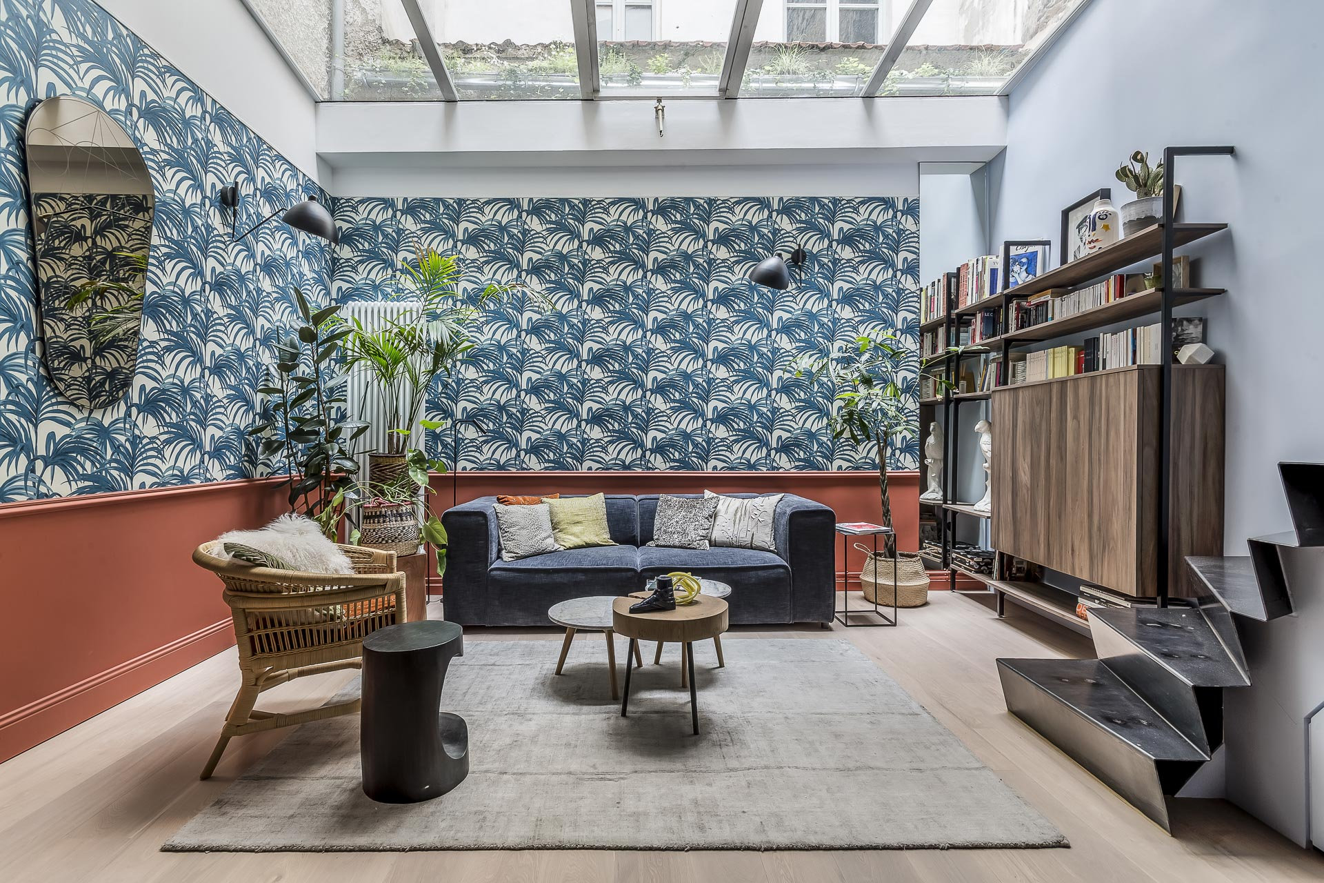 Un salon élégant avec un plafond en verre, un papier peint bleu à feuilles de palmier, un canapé marine avec des coussins, une chaise en osier, des plantes, des étagères et un décor moderne, avec une lumière naturelle et une atmosphère chaleureuse. | 2BeApart Lyon