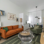 Salon moderne avec un canapé orange, un fauteuil vert, une chaise claire et une table basse ronde. Cuisine et salle à manger en arrière-plan, avec des armoires blanches et une table pour deux personnes. L'art et la décoration ajoutent de la couleur à cet espace lumineux. | 2BeApart Lyon
