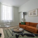 Un salon moderne avec un canapé orange brûlé, une chaise transparente, une table basse ronde en osier et des œuvres d'art encadrées sur des murs blancs. La lumière pénètre par une grande fenêtre aux rideaux diaphanes. Un tapis vert recouvre le sol en bois. | 2BeApart Lyon