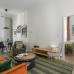Un salon moderne avec un canapé orange, un fauteuil vert et un tapis rayé mène à une salle à manger avec une table ronde et à une chambre lumineuse avec un couvre-lit vert à carreaux, le tout agrémenté d'une décoration contemporaine et d'œuvres d'art sur les murs. | 2BeApart Lyon