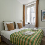 Un lit bien fait avec une couverture à carreaux verts et jaunes, des oreillers bruns et blancs et deux serviettes pliées. La chambre a des murs blancs, une fenêtre avec des rideaux bruns, une petite table de chevet ronde et un tableau encadré. | 2BeApart Lyon