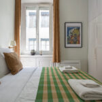 Un lit soigneusement fait avec des oreillers bruns et blancs et une couverture à carreaux verts fait face à une fenêtre aux rideaux dorés. Deux serviettes pliées sont posées sur le lit. Il y a une armoire et des œuvres d'art encadrées au mur. | 2BeApart Lyon
