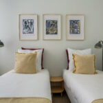 Une chambre bien rangée avec deux lits simples, chacun avec une literie blanche, un coussin jaune et une table de nuit en bois partagée entre eux. Au-dessus des lits se trouvent trois œuvres d'art abstraites encadrées sur un mur clair. | 2BeApart Lyon