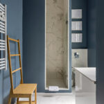 Salle de bains moderne avec des murs bleus, une chaise en bois, un porte-serviettes chauffant, une douche étroite en verre avec des carreaux de marbre, une baignoire blanche et trois œuvres d'art encadrées au mur. Des carreaux de sol blancs complètent l'ensemble. | 2BeApart Lyon