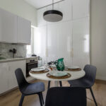 Cuisine moderne avec meubles blancs, dosseret en marbre et sol en bois. Une table ronde est dressée pour quatre personnes avec des chaises noires, des assiettes blanches, des verres, une carafe d'eau verte et des sets de table tissés. Une lampe suspendue noire est accrochée au-dessus. | 2BeApart Lyon