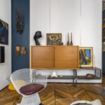 Un salon moderne avec un mélange d'œuvres d'art sur des murs blancs et bleu foncé, une télévision, un meuble en bois, une chaise en fil de fer avec un coussin rouge, un canapé gris et un sol à motifs géométriques en bois. | 2BeApart Lyon