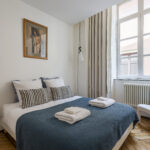 Une chambre confortable avec un lit double, une couverture bleue, des oreillers rayés, des serviettes pliées, une lampe noire, une chaise blanche, une grande fenêtre avec des rideaux et un portrait encadré sur le mur. | 2BeApart Lyon