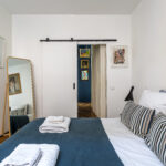 Une chambre moderne avec un lit bien fait, des oreillers rayés et des serviettes pliées, un grand miroir, des murs blancs décorés d'œuvres d'art et une porte coulissante révélant d'autres œuvres d'art dans la pièce adjacente. | 2BeApart Lyon