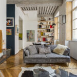 Un salon élégant avec un canapé gris, des coussins rayés, du parquet, des bibliothèques, des œuvres d'art sur les murs, de grandes fenêtres et un éclairage moderne. Le décor mêle des éléments du milieu du siècle et des éléments contemporains. | 2BeApart Lyon