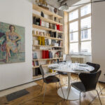 Une petite salle à manger moderne avec une table ronde blanche, quatre chaises foncées, des assiettes et des verres. Derrière, une haute étagère contient des livres et des objets de décoration. Un portrait coloré est accroché au mur de gauche ; de grandes fenêtres laissent entrer la lumière naturelle. | 2BeApart Lyon