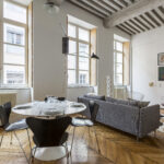 Un salon et une salle à manger modernes avec de grandes fenêtres, des planchers en bois, une table ronde pour quatre personnes, un canapé gris, des chaises contemporaines, des œuvres d'art murales et des poutres apparentes au plafond. L'espace est baigné de lumière naturelle. | 2BeApart Lyon