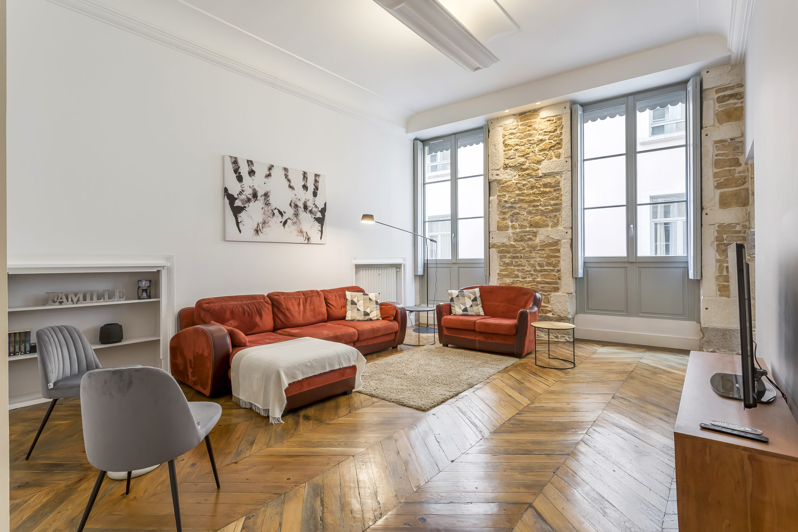 Un salon lumineux avec un sol en bois à chevrons, un canapé rouge avec une couverture, un fauteuil, une chaise grise, de grandes fenêtres, des murs en pierres apparentes, une télévision et une étagère avec des livres et des objets de décoration. | 2BeApart Lyon