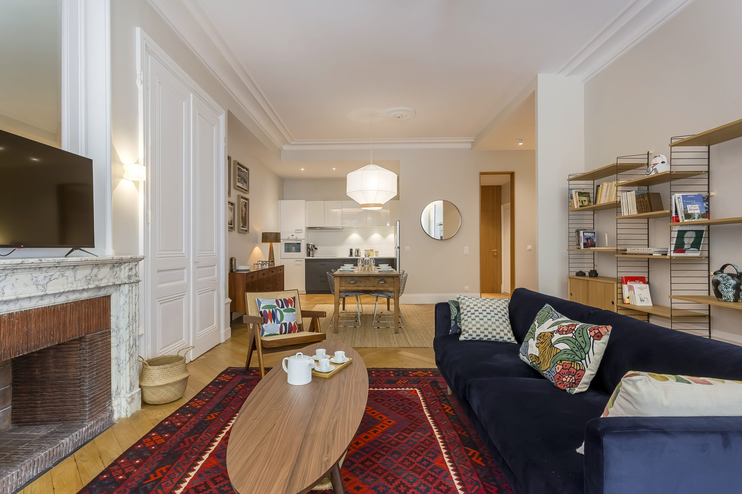 Regulations 5 Un salon moderne avec un canapé bleu foncé, des coussins à motifs, une table basse en bois avec un service à thé, un tapis rouge, une cheminée, une télévision, des étagères, une cuisine ouverte et une salle à manger avec un miroir rond au mur. | 2BeApart Lyon