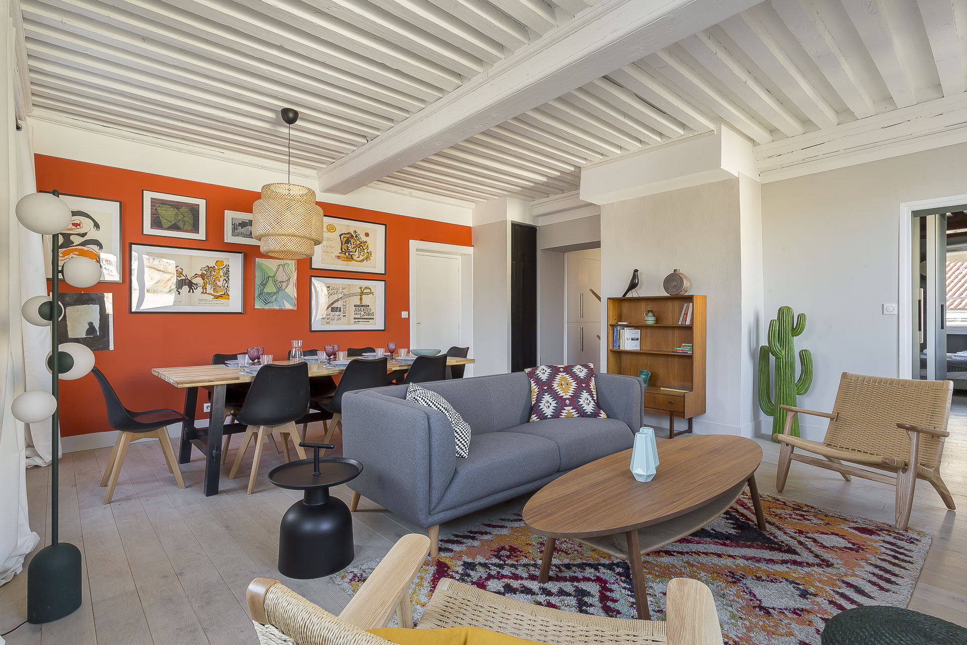 Regulations 8 Un salon et une salle à manger modernes et colorés avec un canapé gris, des chaises en bois, une table basse ronde et une table à manger pour six personnes. La pièce est agrémentée d'un mur d'accent rouge, d'œuvres d'art encadrées et d'objets décoratifs. | 2BeApart Lyon