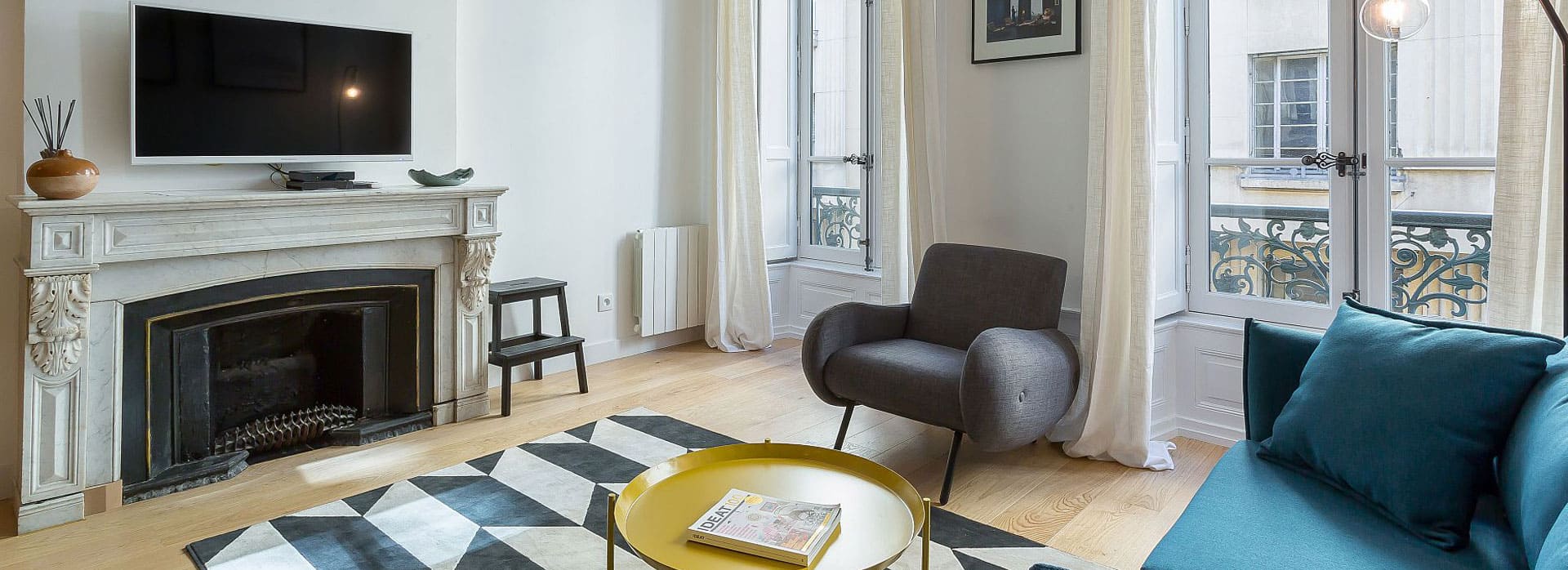 Page d'accueil 4 Un salon élégant avec un canapé en cuir marron, deux fauteuils à carreaux, une télévision murale, des étagères décoratives et de grandes fenêtres qui laissent entrer la lumière naturelle. Les murs sont gris et ornés d'une tête de taureau décorative. | 2BeApart Lyon