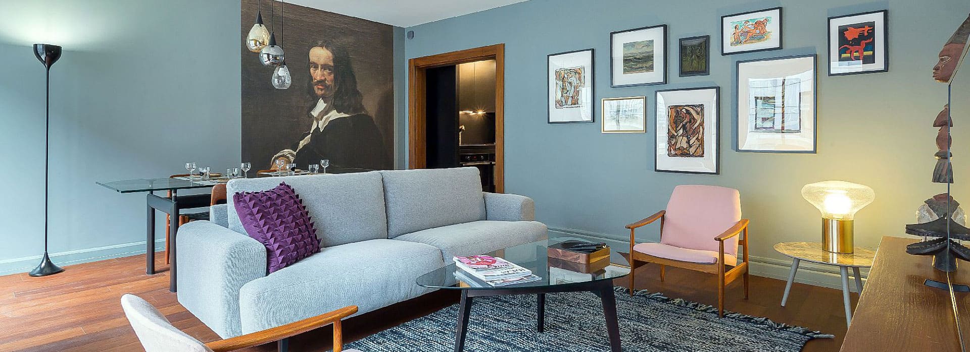 Page d'accueil 3 Salon moderne avec des murs bleu-gris, un sol en bois, un canapé gris, deux fauteuils roses, un tapis noir, des œuvres d'art murales, un lampadaire et un coin repas avec un grand portrait classique en arrière-plan. | 2BeApart Lyon