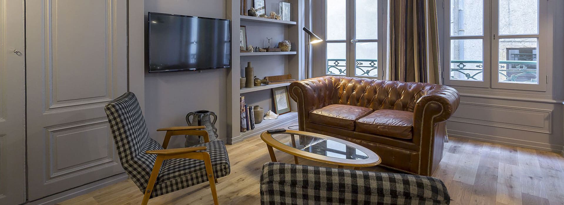 Page d'accueil 5 Salon lumineux avec de hautes fenêtres, des murs blancs, une cheminée en marbre, une télévision murale, un canapé bleu, un fauteuil gris, une table basse ronde jaune, un tapis géométrique et des rideaux transparents laissant entrer la lumière naturelle. | 2BeApart Lyon