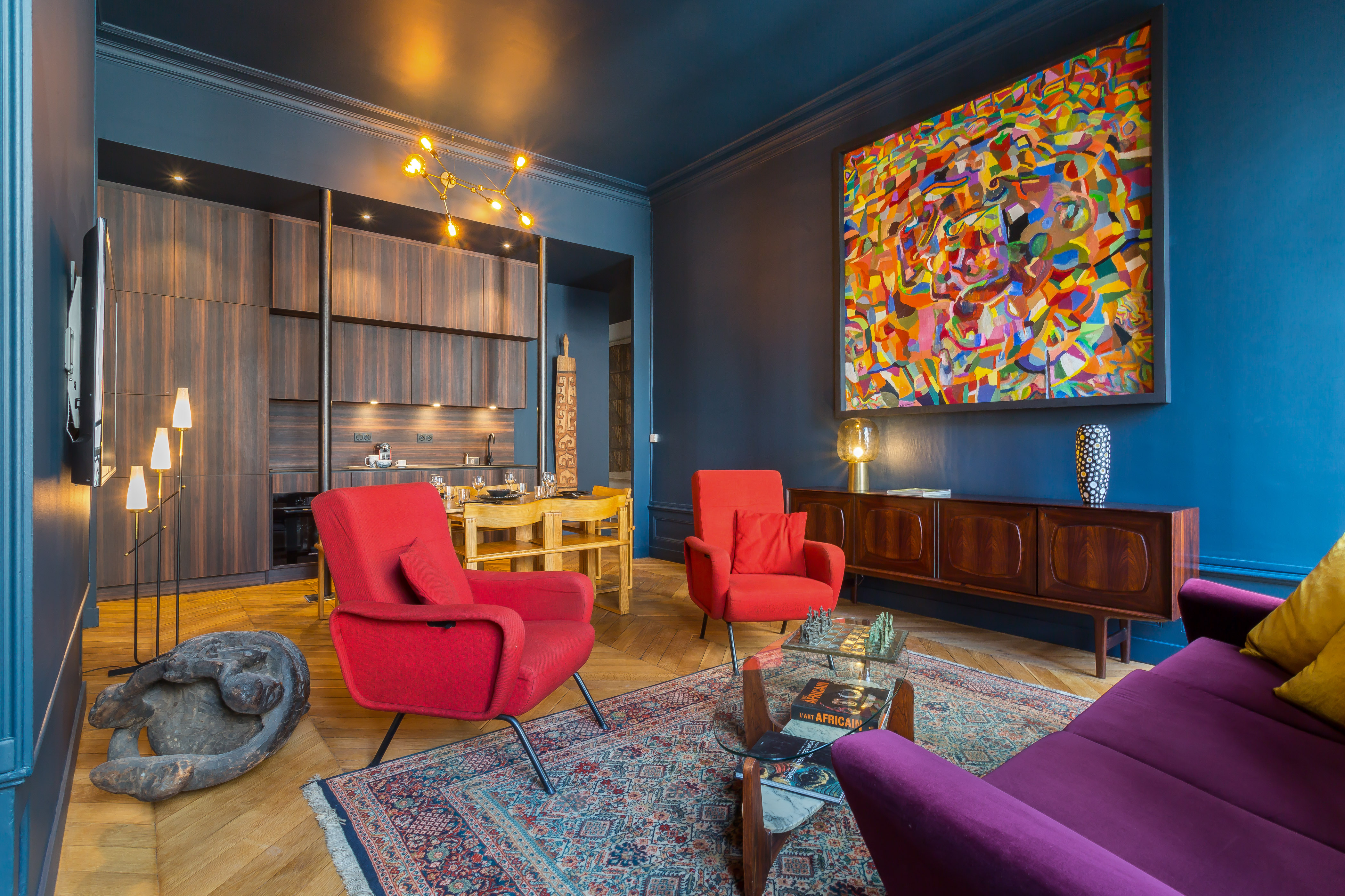 Salon de l’appartement Blue Dream, avec murs bleus, fauteuils rouges et cuisine ouverte.