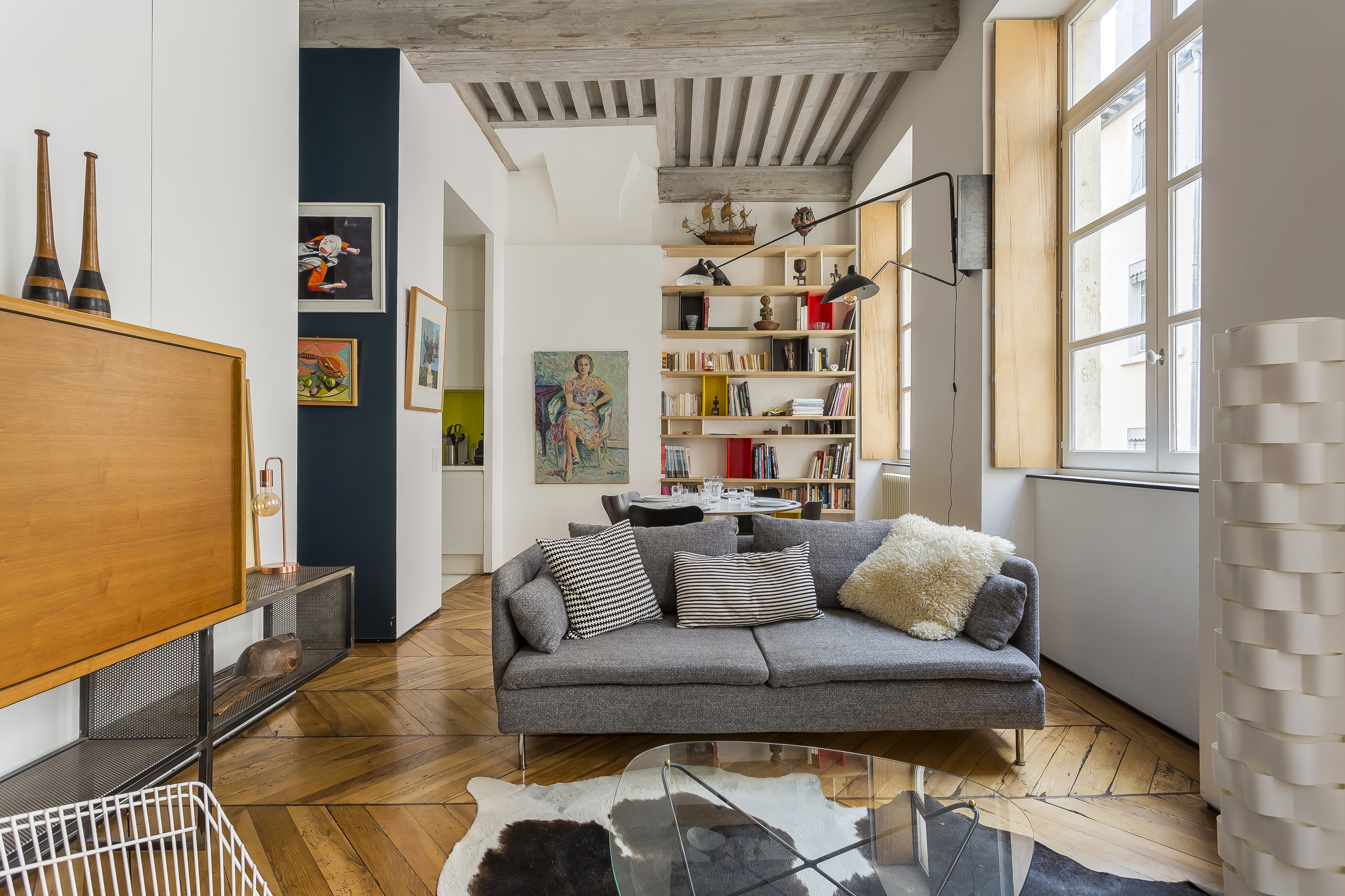 Salon de l’appartement Polycarpe, avec canapé gris, bibliothèque et poutres apparentes.