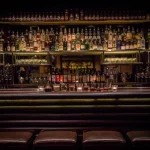 Un bar faiblement éclairé avec des étagères pleines de bouteilles d'alcool et de verres derrière le comptoir, plusieurs bougies sur le bar et des tabourets en cuir alignés devant - un cadre parfait pour les propriétaires exigeants à la recherche d'une conciergerie haut de gamme. | 2BeApart Lyon