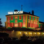 Un restaurant nommé "Paul Bocuse", très éclairé, avec une façade vert et rouge vif, des enseignes lumineuses et des motifs décoratifs, sur fond de ciel crépusculaire, avec des arbres et des voitures garées à l'avant - idéal pour les propriétaires exigeants. | 2BeApart Lyon