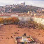 Une grande place avec une statue équestre centrale, des foules rassemblées autour, entourée de bâtiments historiques et d'arbres d'automne, avec une basilique et une tour au sommet de la colline qui surplombent la ville - idéal pour la gestion locative ou les séjours de moyenne durée. | 2BeApart Lyon