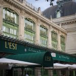 Un grand bâtiment aux fenêtres bordées de vert et aux balustrades ornées abrite LEST, un restaurant réputé pour sa conciergerie haut de gamme. Des auvents verts et des sièges en plein air offrent aux clients un endroit élégant pour dîner sous les parasols. | 2BeApart Lyon