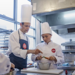 Un chef instructeur en toque et tablier blanc guide un élève chef qui casse un œuf dans un bol dans une cuisine moderne et lumineuse, reflétant l'ambiance d'une conciergerie haut de gamme. D'autres chefs sont partiellement visibles, préparant des aliments sur le comptoir. | 2BeApart Lyon
