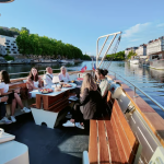 Un groupe de personnes assises autour d'une table sur un bateau à Lyon, dégustant des plats et discutant tout en naviguant le long d'un fleuve bordé d'arbres et de bâtiments sous un ciel bleu clair - un cadre idéal pour une location meublée Lyon en moyenne durée. | 2BeApart Lyon