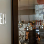 Porte vitrée avec le texte Bistrot Jullien et une vue sur une cuisine moderne à Lyon, où un chef travaille, avec des lampes suspendues et du matériel de cuisine - parfait pour ceux qui recherchent des services de gestion locative ou de conciergerie haut de gamme. | 2BeApart Lyon