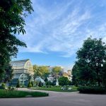Un parc avec un jardin d'hiver en verre, des arbres verdoyants et des gens qui se détendent sur l'herbe sous un ciel bleu avec des nuages ondulants - un endroit invitant pour les propriétaires exigeants qui recherchent le calme, même pour une courte durée. Des bâtiments s'élèvent gracieusement à l'arrière-plan. | 2BeApart Lyon