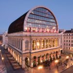 Un grand bâtiment historique avec une extension moderne en dôme de verre est éclairé chaleureusement au crépuscule, sur une place de la ville avec une architecture traditionnelle et des rues légèrement illuminées - idéal pour une gestion locative ou une conciergerie haut de gamme. | 2BeApart Lyon