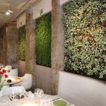 Intérieur d'un restaurant moderne avec des tables et des chaises blanches, des verres à vin posés sur les tables et des murs de jardin verticaux verts entre des piliers en pierre brute. Des fleurs rouges dans un vase décorent une table au premier plan - idéal pour les propriétaires exigeants. | 2BeApart Lyon