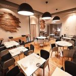 Intérieur de restaurant moderne avec des tables blanches, des chaises noires et grises, des sols en bois et de grandes lampes suspendues rondes. Des œuvres d'art murales en bois décorent les murs, créant une atmosphère de restaurant confortable et élégante avec une touche de conciergerie haut de gamme. | 2BeApart Lyon