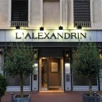 L'extérieur du restaurant L'Alexandrin, idéal pour les propriétaires exigeants, présente une façade moderne avec de grandes fenêtres, deux arbres en pot et une enseigne lumineuse au-dessus de l'entrée. | 2BeApart Lyon