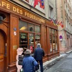Deux personnes se dirigent vers l'entrée du Café des Fédérations, un restaurant traditionnel à l'extérieur en bois dans les rues étroites de Lyon, orné de drapeaux colorés au-dessus - parfait pour les propriétaires exigeants à la recherche d'un charme authentique. | 2BeApart Lyon