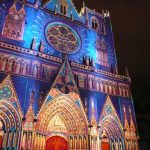 Une cathédrale illuminée la nuit par des lumières bleues, roses et jaunes vibrantes, mettant en valeur son architecture gothique ornée et sa rosace. Une foule s'amasse en contrebas, silhouettée par le spectacle - parfait pour ceux qui profitent des services de location meublée Lyon ou de conciergerie haut de gamme. | 2BeApart Lyon