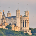 Une grande basilique blanche avec quatre tours et des croix se dresse au sommet d'une colline verdoyante, entourée d'arbres et de bâtiments historiques, sous un ciel doux et nuageux - une vue emblématique près de nombreuses options de location meublée à Lyon. Des oiseaux volent autour de la structure. | 2BeApart Lyon
