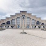 Un grand bâtiment symétrique avec de hautes fenêtres cintrées et un toit en gradins se dresse derrière une place pavée ouverte, dotée d'une haute colonne centrale et de deux fontaines circulaires. Idéal pour les propriétaires exigeants à la recherche d'une conciergerie haut de gamme. Le ciel est couvert. | 2BeApart Lyon