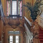 Grand escalier en bois avec sculptures ornementales et tapis rouge, grand vitrail au-dessus, palmier en pot sur un piédestal décoratif - un cadre impressionnant idéal pour la gestion locative, que ce soit pour des séjours de moyenne durée ou de courte durée. Portes à double battant avec verre dépoli en dessous. | 2BeApart Lyon
