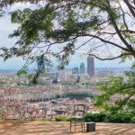 Une chaise et une petite table sont installées sur une plate-forme en bois sous un grand arbre, avec vue sur un paysage urbain panoramique. C'est l'endroit idéal à Lyon pour les propriétaires exigeants à la recherche d'un emplacement meublé avec une vue imprenable. | 2BeApart Lyon
