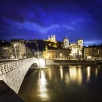 Une vue nocturne d'un pont en pierre au-dessus d'un fleuve à Lyon, en France, avec des bâtiments historiques illuminés - dont la basilique Notre-Dame de Fourvière - sur fond de ciel bleu profond, idéal pour ceux qui recherchent une location meublée Lyon ou une conciergerie haut de gamme. | 2BeApart Lyon