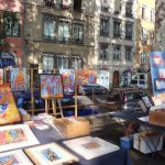 Des peintures et des gravures colorées sont exposées sur des tables et des chevalets lors d'un marché d'art en plein air à Lyon, avec des voitures garées et des immeubles d'habitation à proximité - une scène invitante pour les personnes intéressées par les services de location meublée Lyon ou de gestion locative. | 2BeApart Lyon