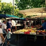 Des acheteurs, dont un enfant et des adultes, parcourent des produits frais sur un marché de producteurs en plein air, sous des arbres feuillus. Cette scène animée reflète le mode de vie dynamique des propriétaires exigeants qui recherchent une location meublée à Lyon. | 2BeApart Lyon